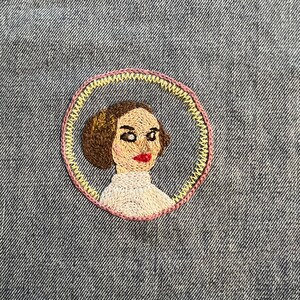 May include: Embroidered patch of a woman with brown hair, wearing a white shirt, with a yellow and pink circle around her.