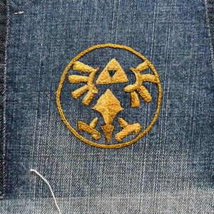 May include: Close-up of a denim patch with a gold embroidered Triforce symbol, a popular design from the Legend of Zelda video game series. The patch has a frayed edge and is sewn onto the denim fabric.