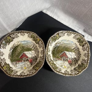 May include: Two ceramic bowls featuring a central illustration of a red covered bridge in a snowy landscape. The rim of each bowl is decorated with a detailed brown and green leaf pattern on a cream background with a scalloped edge design.