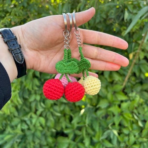 Könnte beinhalten: Ein Schlüsselanhänger mit drei gehäkelten Kirschen in Rot, Rosa und Gelb mit grünen Blättern. Die Kirschen sind an einem silbernen Schlüsselring befestigt.