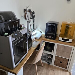 Peut inclure: Un espace de bureau à domicile avec plusieurs imprimantes 3D, dont des modèles Anycubic. L'espace de travail comprend un bureau, une chaise et des meubles de rangement. Diverses bobines de filament sont visibles, ainsi qu'une plante succulente murale.