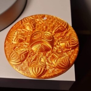 May include: A round, orange-colored decorative item with a series of embossed scarab beetle designs. The central design features a stylized face. The item has a small hole at the top for hanging.