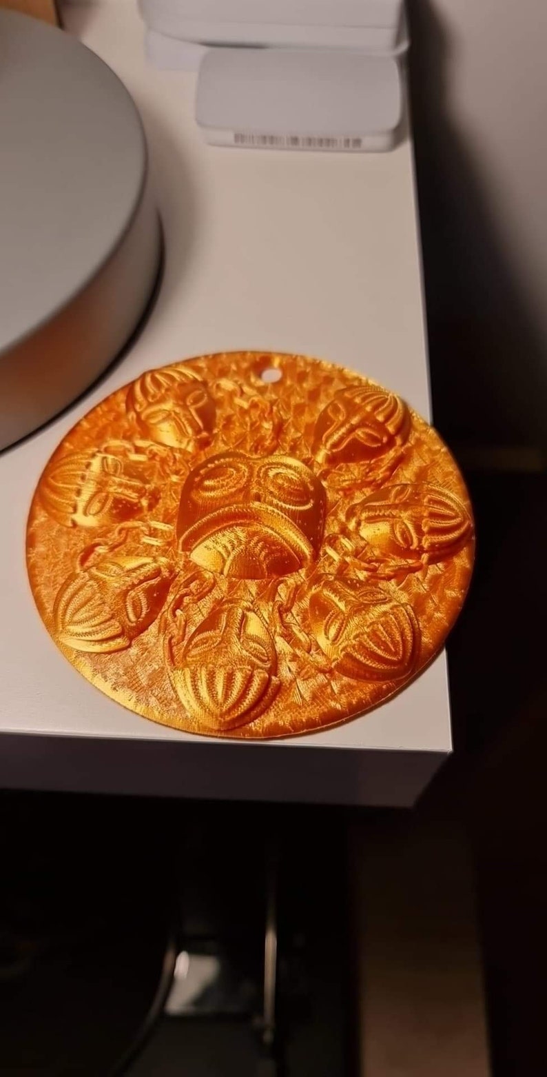 May include: A round, orange-colored decorative item with a series of embossed scarab beetle designs. The central design features a stylized face. The item has a small hole at the top for hanging.