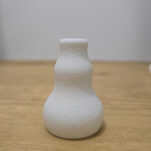 May include: A small, white, textured vase with a bulbous base and a narrow neck. The vase has a matte finish and sits on a wooden surface. The overall shape is reminiscent of a gourd.