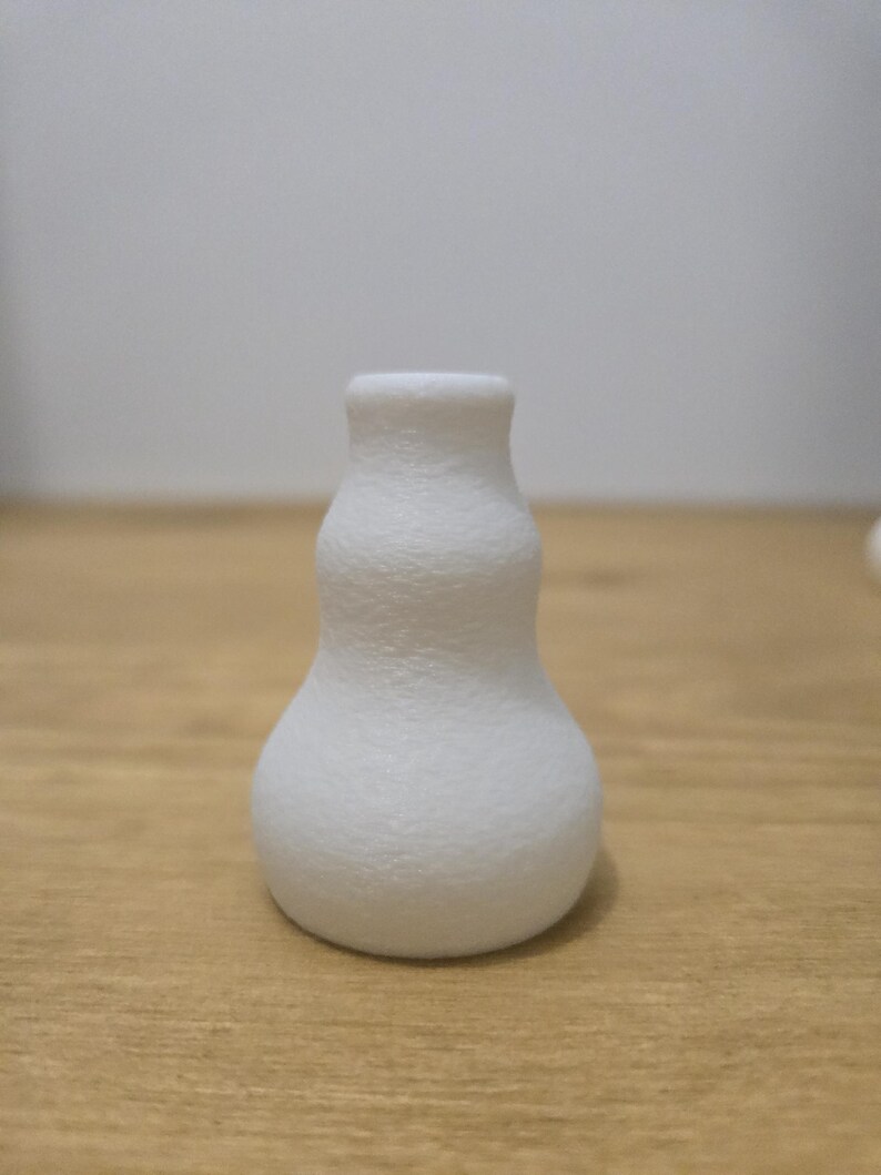 May include: A small, white, textured vase with a bulbous base and a narrow neck. The vase has a matte finish and sits on a wooden surface. The overall shape is reminiscent of a gourd.