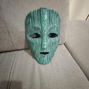 May include: A green mask with a textured, wood-like appearance. The mask has eye and mouth openings, and a detailed design on the forehead. The mask is resting on a gray surface.
