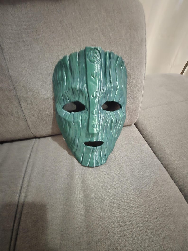 May include: A green mask with a textured, wood-like appearance. The mask has eye and mouth openings, and a detailed design on the forehead. The mask is resting on a gray surface.