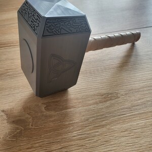 May include: A gray, geometric hammer with a wrapped handle. The hammer head features engraved patterns and a circular emblem. The handle is wrapped in a light brown material. The hammer is resting on a wooden surface.
