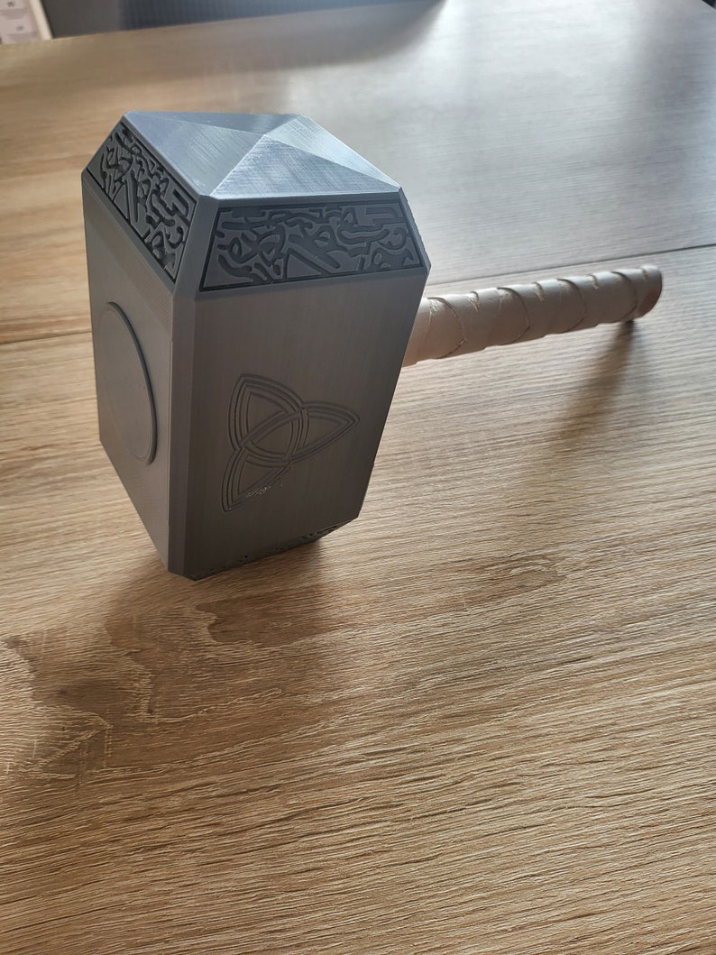 May include: A gray, geometric hammer with a wrapped handle. The hammer head features engraved patterns and a circular emblem. The handle is wrapped in a light brown material. The hammer is resting on a wooden surface.