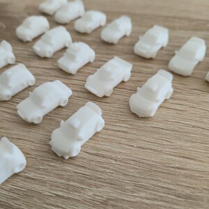 May include: A collection of small, white toy cars scattered on a light brown wooden surface. The miniature vehicles are uniformly white, with visible details like windows and roofs. The cars are arranged in a seemingly random pattern.