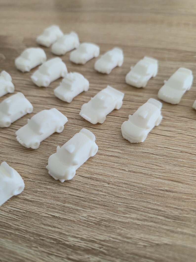 May include: A collection of small, white toy cars scattered on a light brown wooden surface. The miniature vehicles are uniformly white, with visible details like windows and roofs. The cars are arranged in a seemingly random pattern.