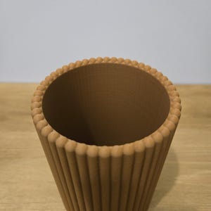 May include: A brown, decorative cup-shaped container with a ridged exterior and a beaded rim. The interior is smooth and the container is set on a wooden surface. The design is reminiscent of a cupcake liner.