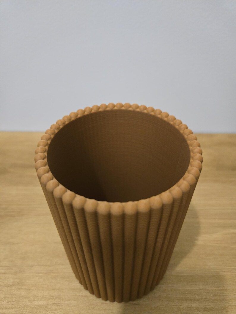 May include: A brown, decorative cup-shaped container with a ridged exterior and a beaded rim. The interior is smooth and the container is set on a wooden surface. The design is reminiscent of a cupcake liner.