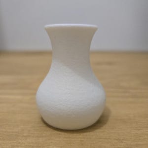 May include: A small, white vase with a textured surface. The vase has a rounded base, a narrow neck, and a flared rim. The vase is approximately 4 inches tall.