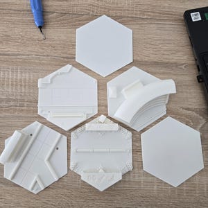 May include: A collection of white, hexagonal 3D-printed objects, some with intricate designs and architectural elements. The objects are arranged on a wooden surface, alongside a blue tool and a laptop.