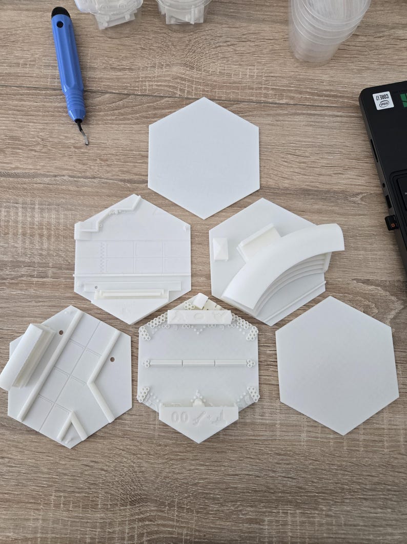 May include: A collection of white, hexagonal 3D-printed objects, some with intricate designs and architectural elements. The objects are arranged on a wooden surface, alongside a blue tool and a laptop.