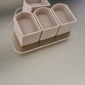 May include: A set of four beige 3D-printed containers with rounded tops and a rectangular base, arranged on a clear, rectangular tray. The containers have a textured interior. The tray sits on a light beige surface.
