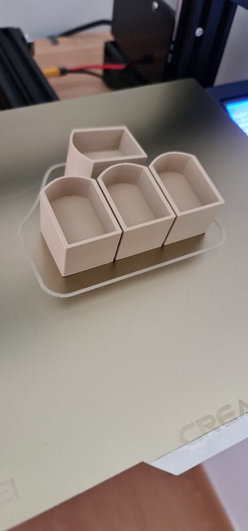 May include: A set of four beige 3D-printed containers with rounded tops and a rectangular base, arranged on a clear, rectangular tray. The containers have a textured interior. The tray sits on a light beige surface.