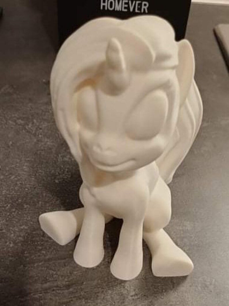 May include: A cream-colored unicorn figurine with a flowing mane and tail, and a single horn. The figurine is in a sitting position, with its front legs extended. The word "HOMEEVER" is visible in the background.