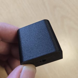 May include: A small, black, rectangular object with a textured surface. The object has a slightly angled top and a small hole on one side. The item is held between fingers against a light wood background.