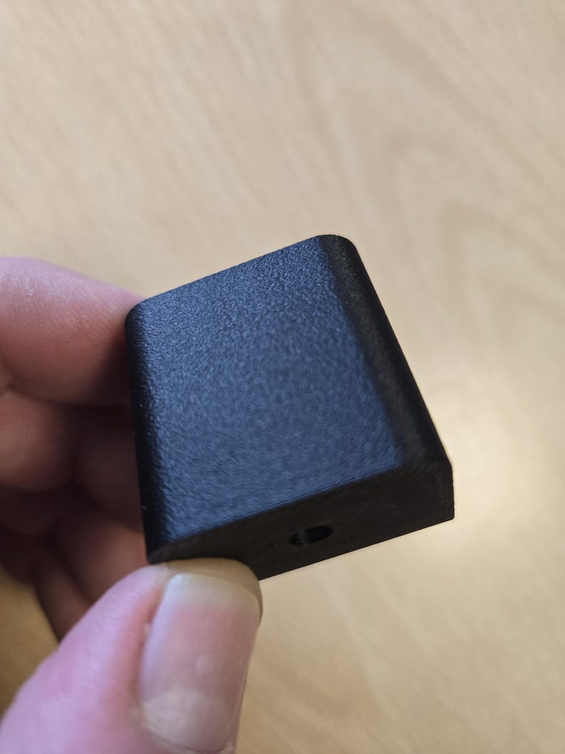 May include: A small, black, rectangular object with a textured surface. The object has a slightly angled top and a small hole on one side. The item is held between fingers against a light wood background.