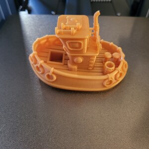 May include: A 3D-printed toy boat in a light orange color. The boat features a cabin with windows, a smokestack, and decorative elements along the sides. Two small barrels are visible on the deck.