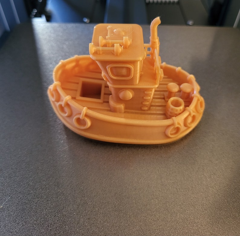 May include: A 3D-printed toy boat in a light orange color. The boat features a cabin with windows, a smokestack, and decorative elements along the sides. Two small barrels are visible on the deck.