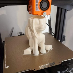 May include: A 3D-printed, off-white pony figurine is being created by a 3D printer. The pony is in a sitting position with its head tilted slightly. The printer head is orange and labeled "CAUTION HOT".