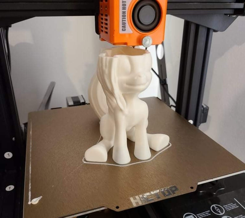 May include: A 3D-printed, off-white pony figurine is being created by a 3D printer. The pony is in a sitting position with its head tilted slightly. The printer head is orange and labeled "CAUTION HOT".
