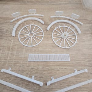 May include: White plastic model kit components, including two wagon wheels, curved arches, rectangular pieces, and support beams. The parts are arranged on a light wood-grain surface, suggesting a craft or hobby project.