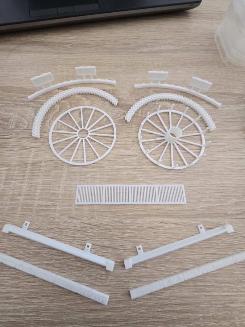 May include: White plastic model kit components, including two wagon wheels, curved arches, rectangular pieces, and support beams. The parts are arranged on a light wood-grain surface, suggesting a craft or hobby project.