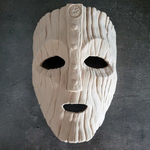 May include: A light-colored, textured mask with a carved wooden appearance. The mask has large eye openings, a small mouth opening, and a central vertical panel with three circular details. A circular emblem is at the top.
