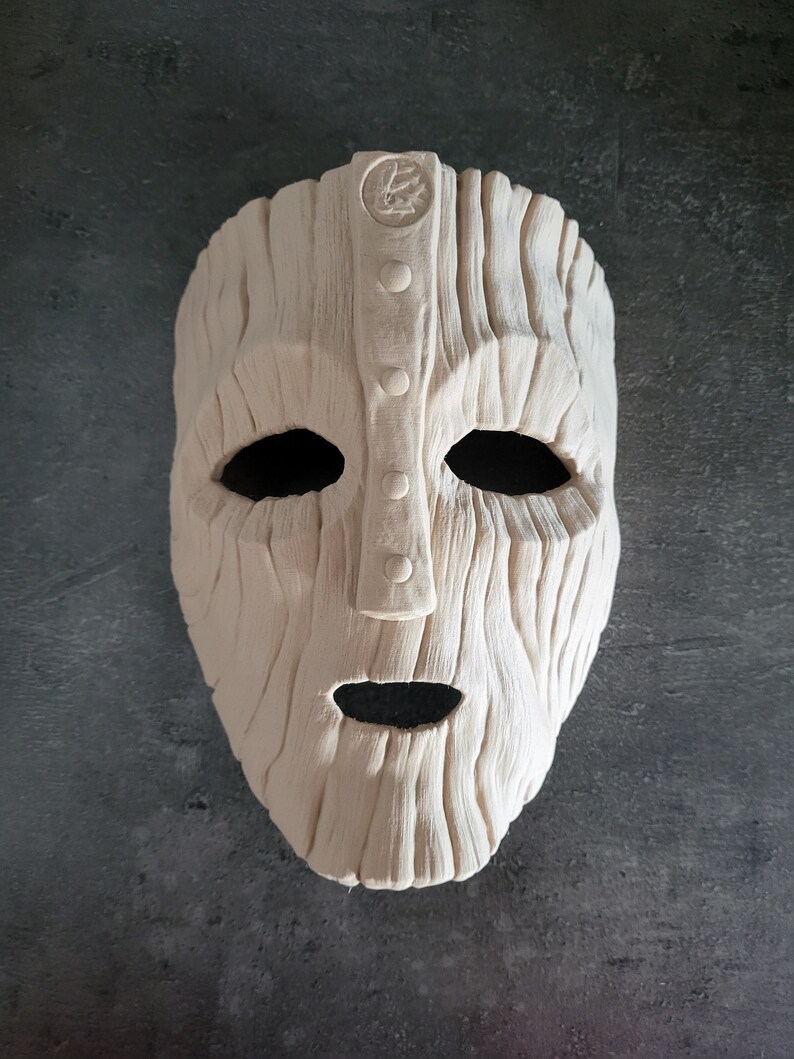 May include: A light-colored, textured mask with a carved wooden appearance. The mask has large eye openings, a small mouth opening, and a central vertical panel with three circular details. A circular emblem is at the top.