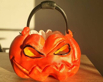May include: Orange plastic Halloween pumpkin bucket with a black handle and a scary face. The bucket has yellow eyes and a wide, toothy grin.