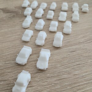May include: A collection of small, white toy cars arranged on a light brown wooden surface. The cars are uniformly shaped, with a simple design. The arrangement suggests a playful or decorative purpose, possibly for a child's toy set or craft project.