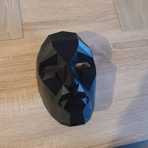 May include: A black, geometric-shaped mask with a human face design. The mask has a polygonal structure with eye and mouth openings. The mask is made of a solid, opaque material and is resting on a light-colored surface.