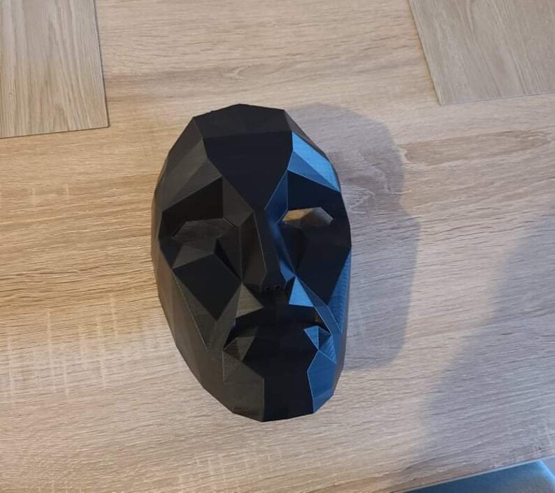 May include: A black, geometric-shaped mask with a human face design. The mask has a polygonal structure with eye and mouth openings. The mask is made of a solid, opaque material and is resting on a light-colored surface.
