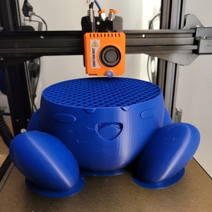May include: A blue 3D-printed character is being created by a 3D printer. The character has a round body with large feet and a smiling face. The printer head is orange and has the text "CAUTION HOT".