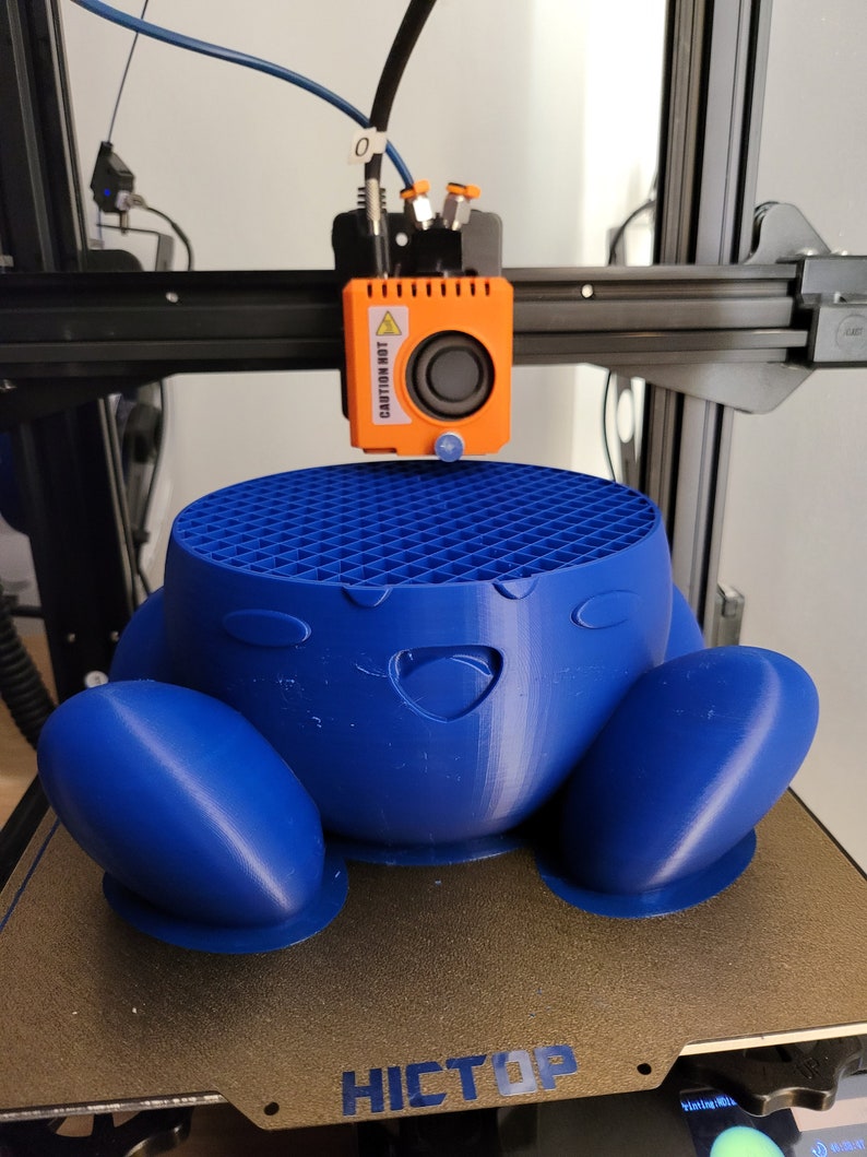 May include: A blue 3D-printed character is being created by a 3D printer. The character has a round body with large feet and a smiling face. The printer head is orange and has the text "CAUTION HOT".