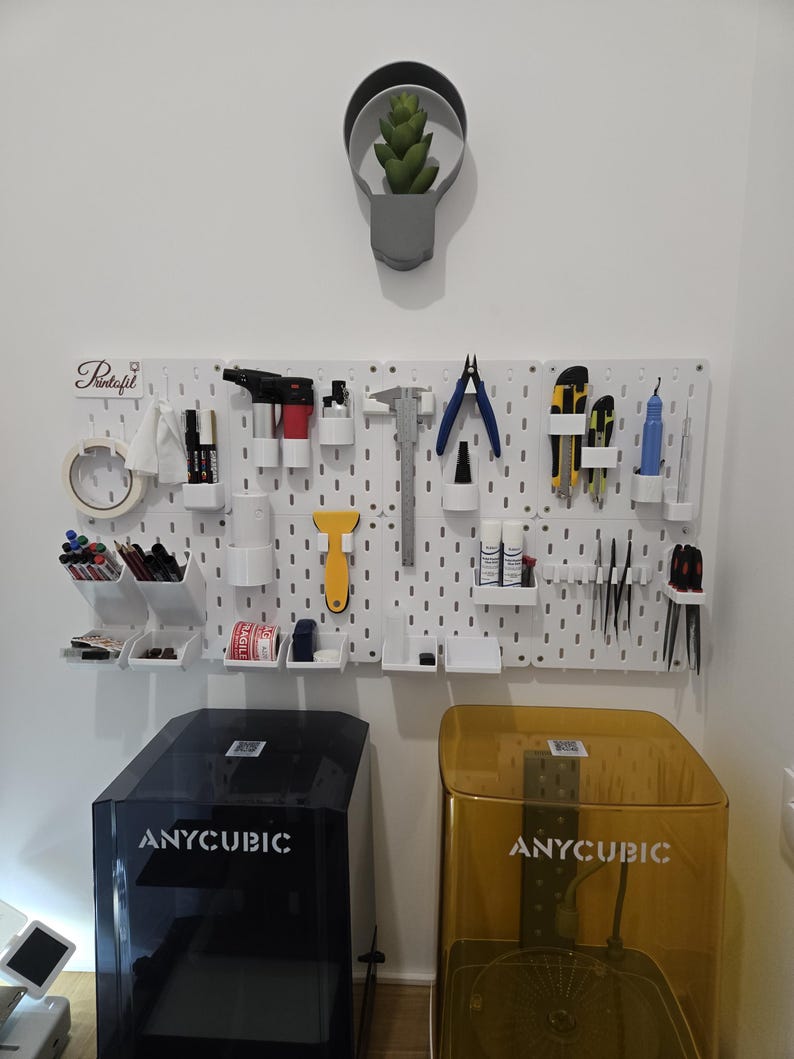 May include: A white pegboard with various tools and supplies, including pliers, markers, and tape. Below are two Anycubic 3D printers, one black and one gold. A small plant in a gray pot hangs above the pegboard.