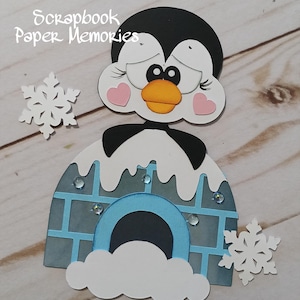 May include: A papercraft penguin with a black head, white face, and orange beak, set against a blue igloo. The penguin has pink heart-shaped cheeks and is surrounded by white snowflakes. The text "Scrapbook Paper Memories" is at the top.