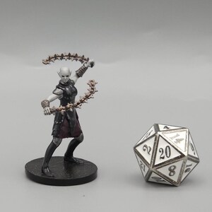 May include: A miniature fantasy figure of a female elf in a black and burgundy outfit, wielding two spiked chains. A twenty-sided die with white faces and black numbers is next to the figure. The figure is on a black base.