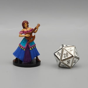 Pode incluir: Uma figura em miniatura de uma bardo feminina tocando um alaúde. Ela está vestindo um vestido azul e roxo com detalhes dourados. A figura está em pé sobre uma base preta. Um dado branco de 20 faces está no fundo.