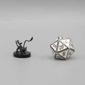 May include: A black miniature figurine of a creature with a long, curled tail. The creature has a cat-like face and is standing on a black base. A white 20-sided die is in the background.