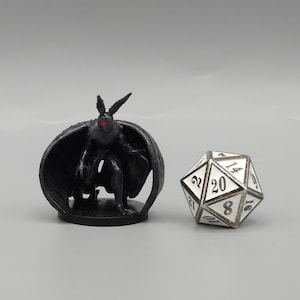 May include: A black miniature figure of a creature with bat-like wings and red eyes. The figure is standing on a black base and is posed in a crouching position. A white 20-sided die is in the background.