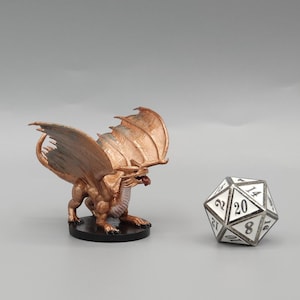 Könnte beinhalten: Eine bronzefarbene Drachenfigur mit ausgebreiteten Flügeln, auf einem schwarzen Sockel. Ein zwanzigseitiger Würfel mit weißen Flächen und schwarzen Zahlen befindet sich neben dem Drachen. Die Flügel des Drachens sind in einem helleren Bronzeton gehalten.