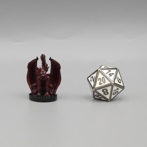 May include: A red plastic miniature dragon figurine with wings spread, standing on a black base. A white 20-sided die with black numbers is also in the image.