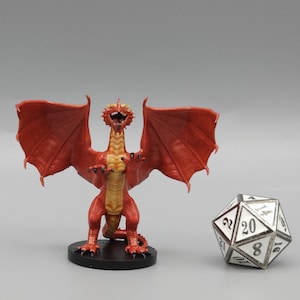 Puede incluir: Una miniatura de dragón rojo con detalles dorados, de pie sobre una base negra. El dragón tiene las alas extendidas y la boca abierta en un rugido. La miniatura mide aproximadamente 5 cm de alto y está hecha de plástico.
