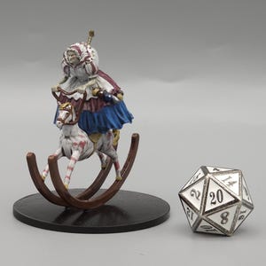 May include: A miniature figurine of a person riding a rocking horse, with a multi-sided die next to it. The rocking horse is brown with a black base. The figurine is wearing a blue skirt and a red cape.