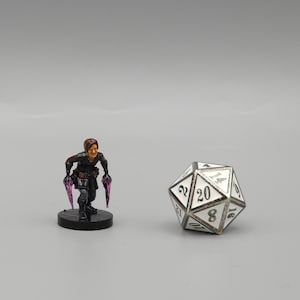 Puede incluir: Una figura miniatura de un personaje femenino con ropa negra que sostiene dos dagas moradas. La figura está de pie sobre una base negra. Un dado blanco de 20 caras está en el fondo.
